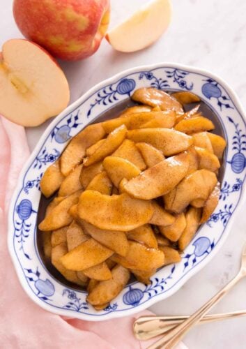 An oval platter of fried apples beside a cut apple, pink linen napkin, and two forks.