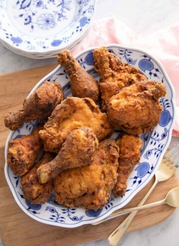 Overhead view of of a platter of fried chicken on a wooden board with two forks and a stack of plates beside it.