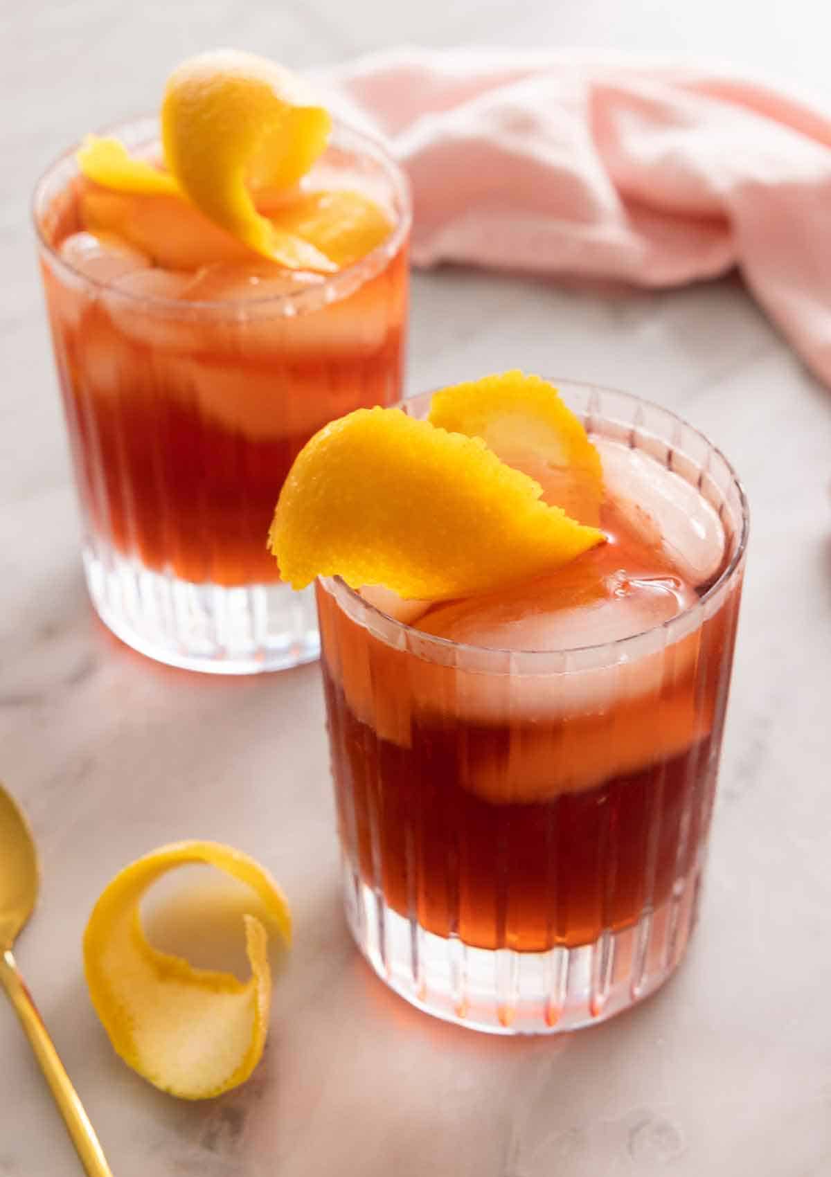 Two glasses of Negroni cocktails with orange peels as garnish.