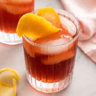 A glass of Negroni with orange peel garnish and ice cubes on a counter with a pink linen napkin and an extra curled orange peel.