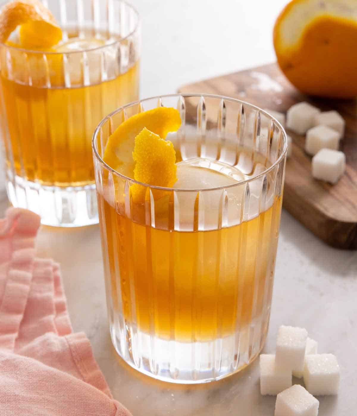 Two glasses of old fashioned cocktails with orange peels in the glass and some sugar cubes on the side.