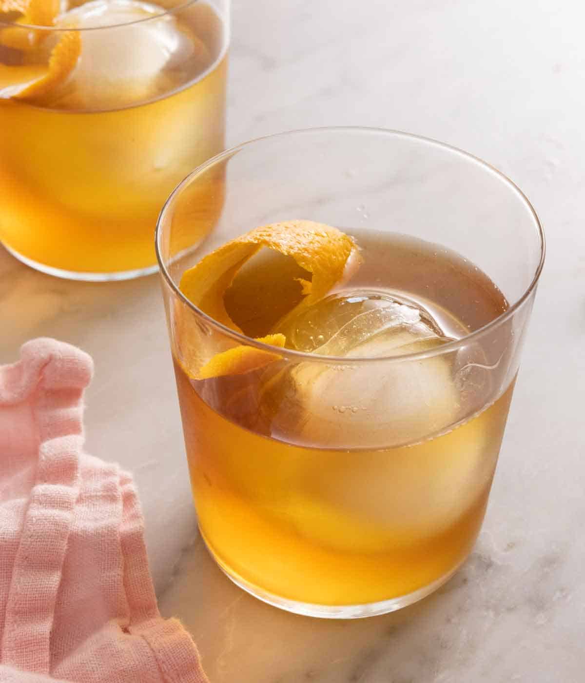 A glass of old fashioned with a circular ice cube and orange peel inside. A second drink in the background.