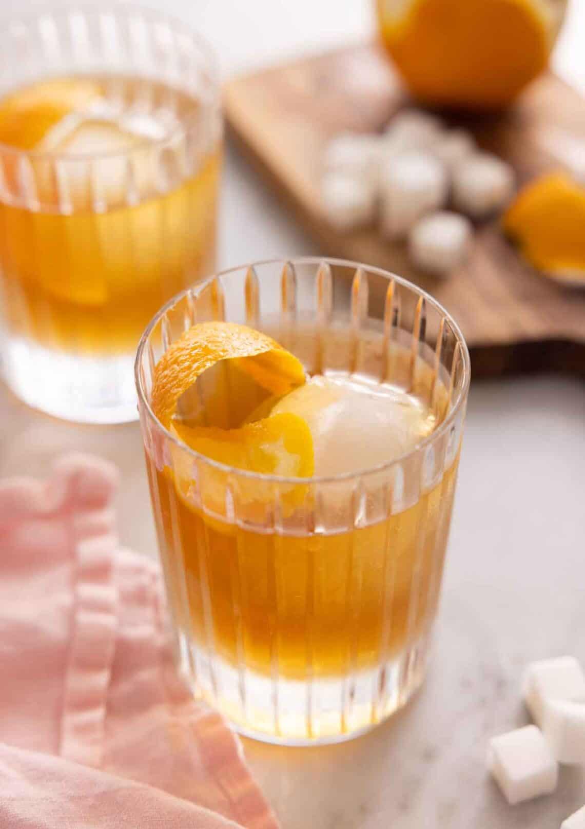 Two glasses of old fashioned with the ingredients in the background, out of focus.