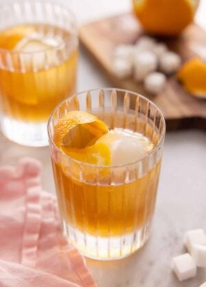 Two glasses of old fashioned with the ingredients in the background, out of focus.