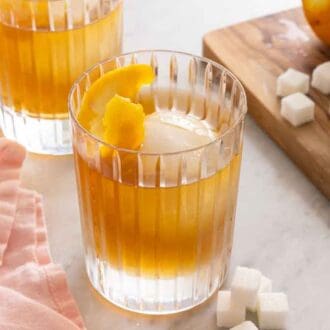 One of two glasses of old fashioned cocktails beside a cutting board with orange peels and sugar cubes.