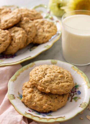 A plate with two peanut butter oatmeal cookies in front of a glass of milk and a platter of more cookies.