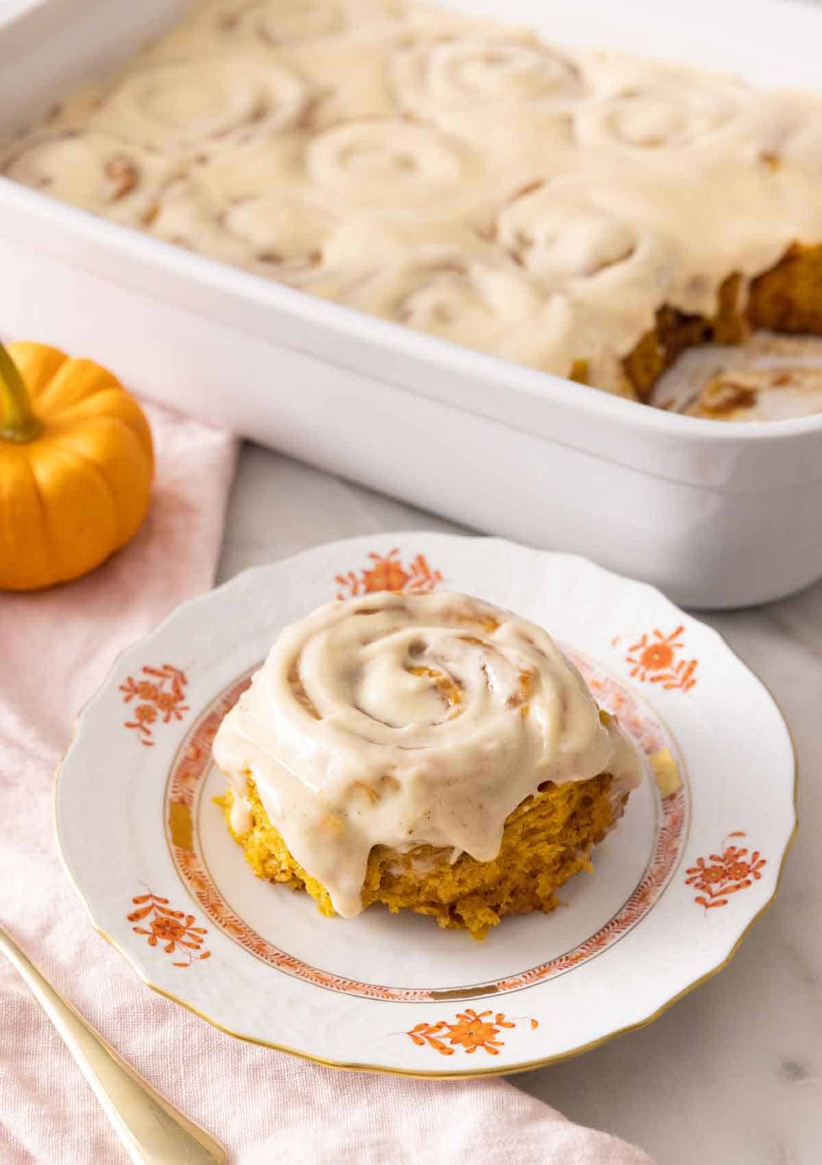 A plate with a single pumpkin cinnamon roll with cream cheese frosting on top in front of a baking pan with the rest.