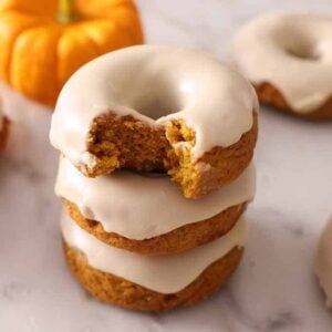 A stack of three glazed pumpkin donuts with the top on with a bite taken out. A few more glazed donuts scattered around.