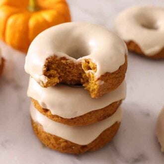 A stack of three glazed pumpkin donuts with the top on with a bite taken out. A few more glazed donuts scattered around.