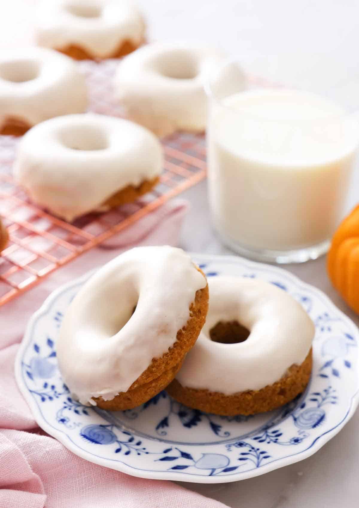 Two glazed pumpkin donuts with one propped against the other on a plate in front of a glass of milk.