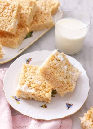 A plate with two squares of rice crispy treats in front of a glass of milk and a platter with more in the back.