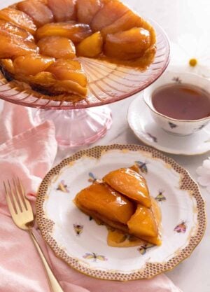 A slice of tarte Tatin on a plate in front of a cake stand with the rest of the Tatin behind it.