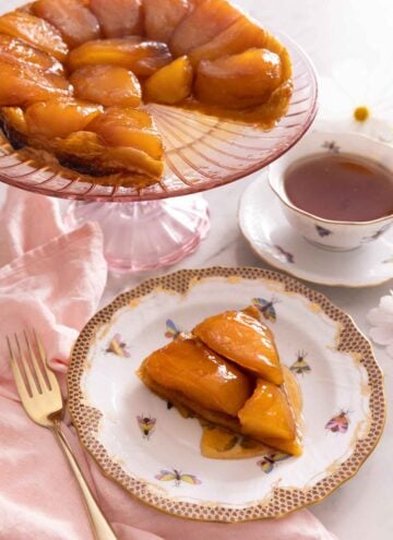A slice of tarte Tatin on a plate in front of a cake stand with the rest of the Tatin behind it.