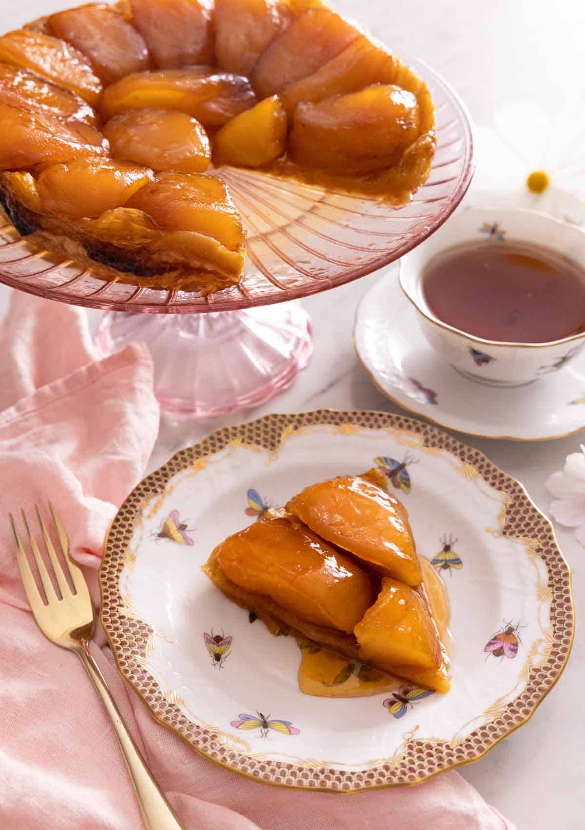 A slice of tarte Tatin on a plate in front of a cake stand with the rest of the Tatin behind it.