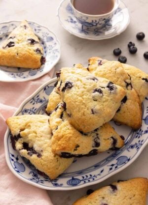 An oval platter of blueberry scones in front of a cup of tea and scattered blueberries on the counter.