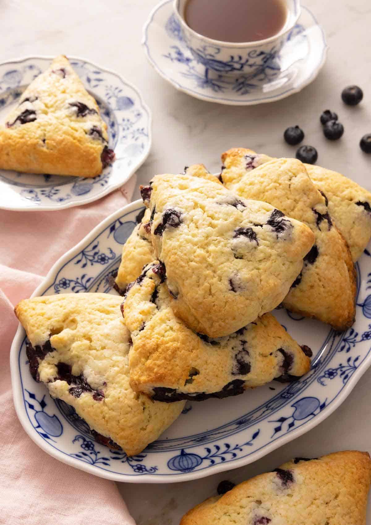 An oval platter of blueberry scones in front of a cup of tea and scattered blueberries on the counter.