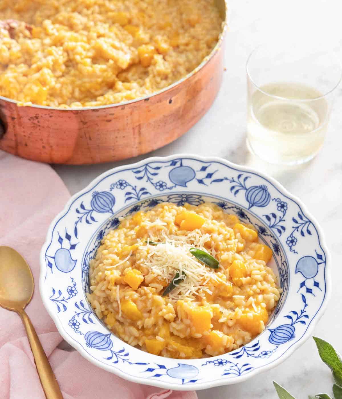 A bowl of butternut squash risotto with shredded cheese sprinkled on top by a glass and pot.