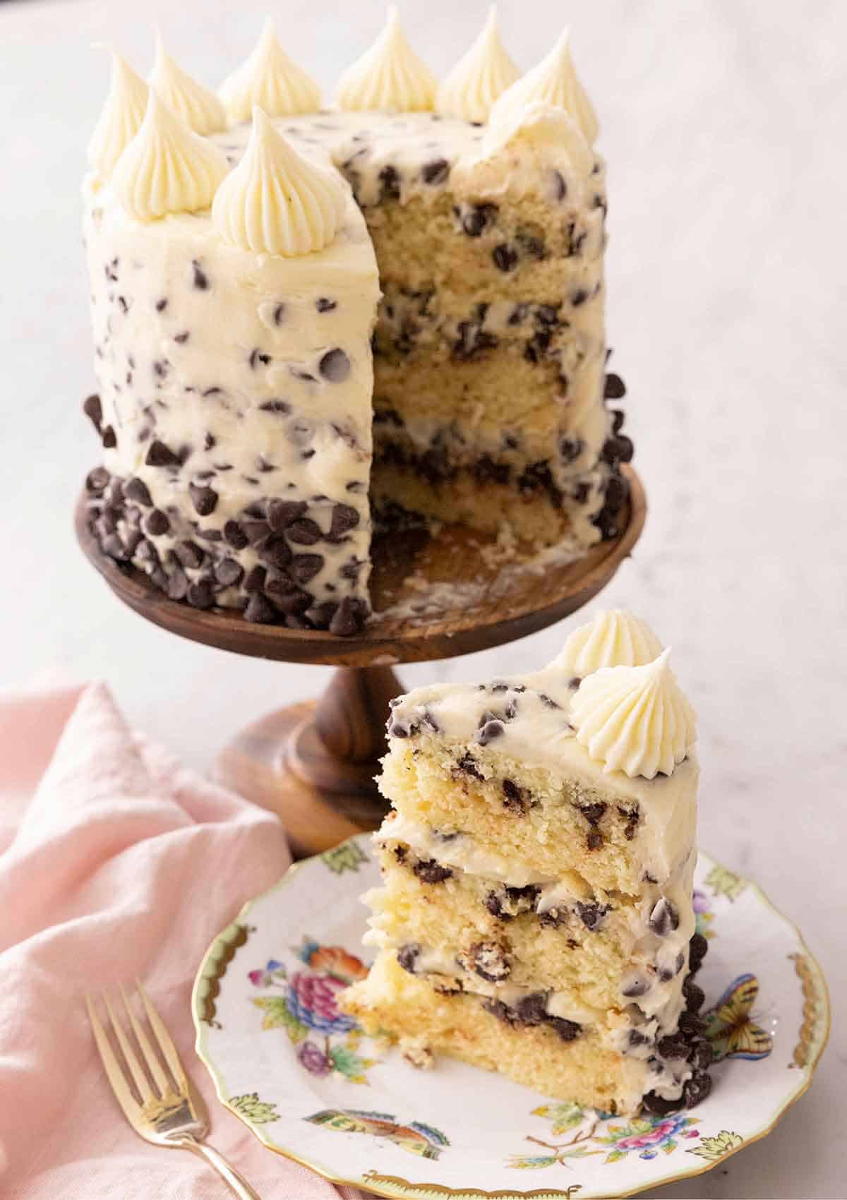 A chocolate chip cake on a cake stand with a slice cut and placed on a plate.