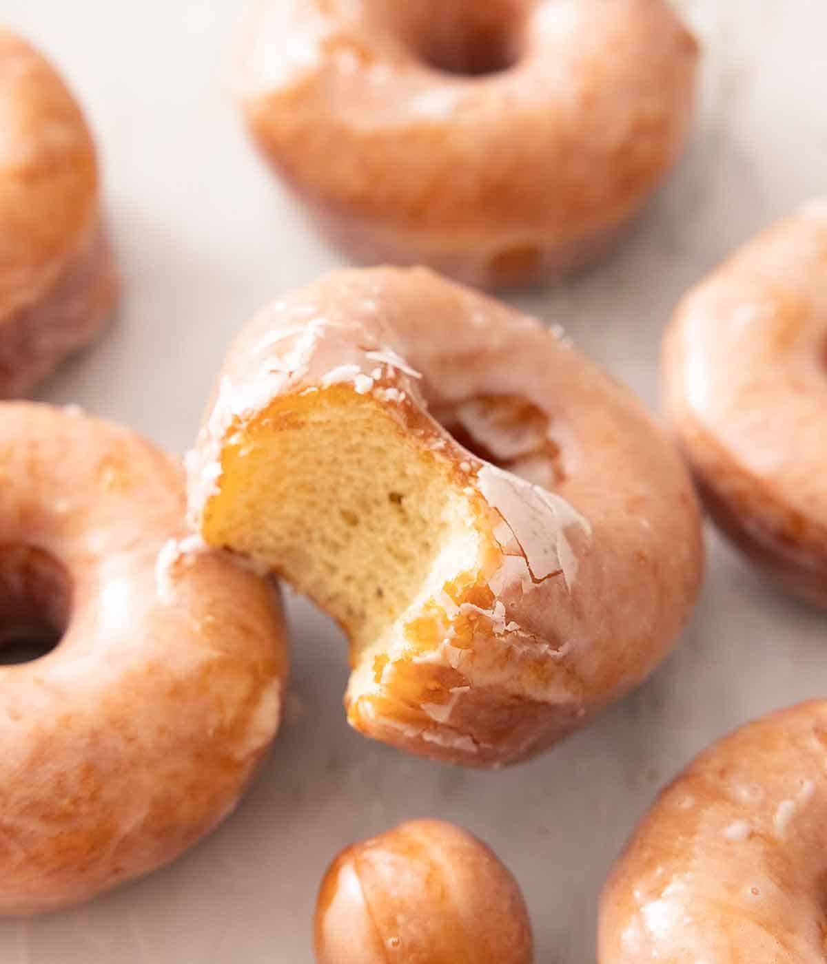 A glazed donut with a bite taken out of it propped on top of more glazed donuts.