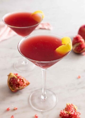 Two glasses of pomegranate martinis with lemon peel garnish.