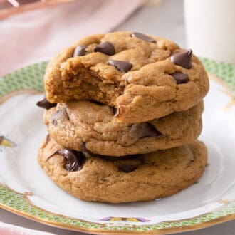A stack of three pumpkin chocolate chip cookies on a plate next to a glass of milk.