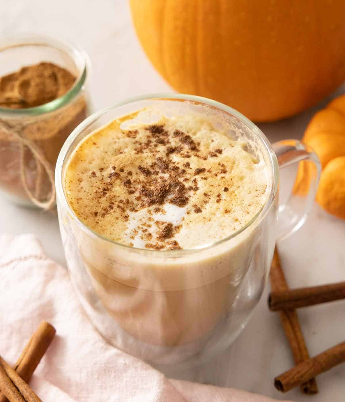 A latte with pumpkin pie spice sprinkled over the foam with cinnamon sticks scattered around and a jar of pumpkin pie spice in the background.