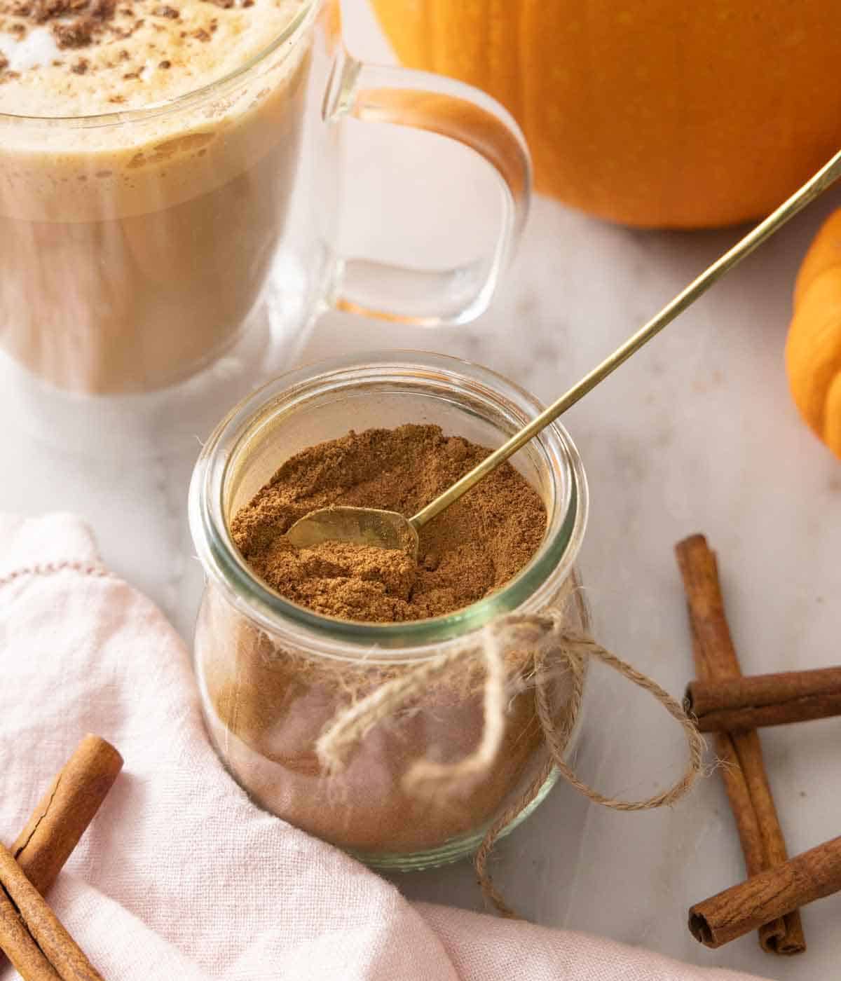 A jar of pumpkin pie spice with a long spoon inside of it. Sticks of cinnamon scattered around.