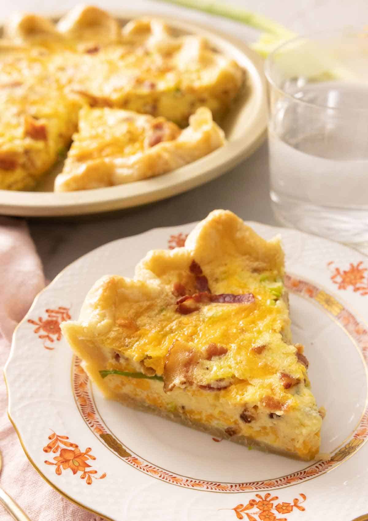 A slice of quiche on a plate in front of the rest of the pie.