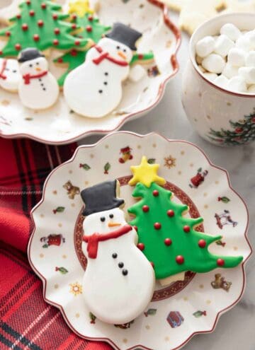A snowman and Christmas tree cookie on a plate decorated with royal icing.