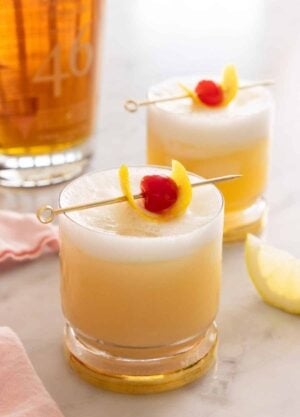 Two glasses of whiskey sour with cocktail picks with lemon peel and maraschino cherry as garnish on top.