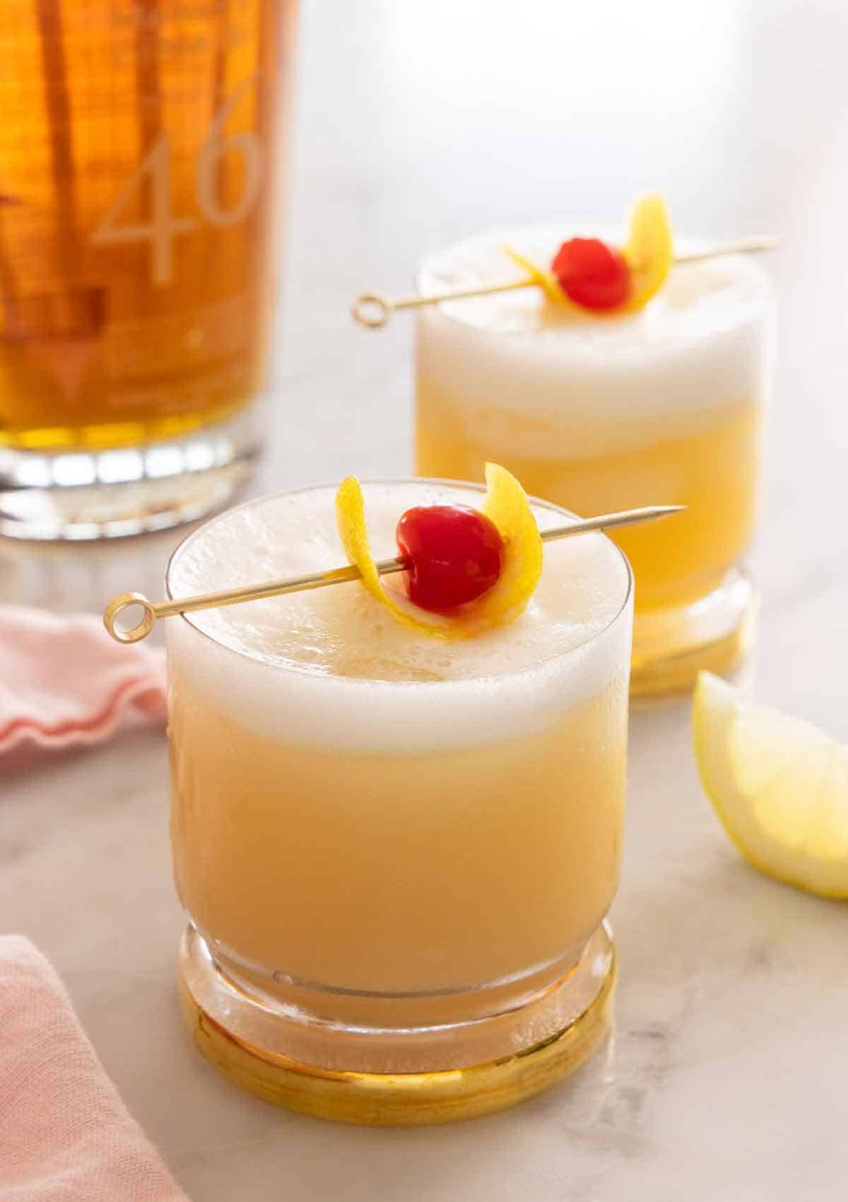 Two glasses of whiskey sour with cocktail picks with lemon peel and maraschino cherry as garnish on top.