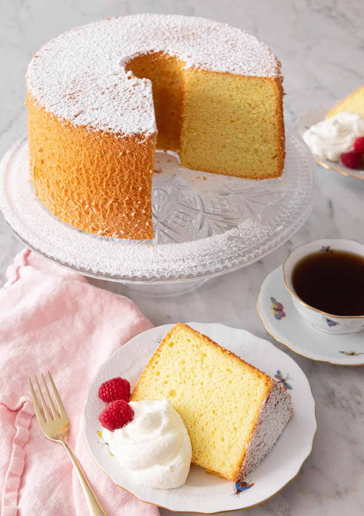 A chiffon cake on a stand, cut with a slice plated in front with whipped cream and berries.