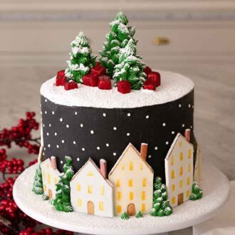 A cake decorated with a snowy top with trees and gifts along with houses pressed to the side.