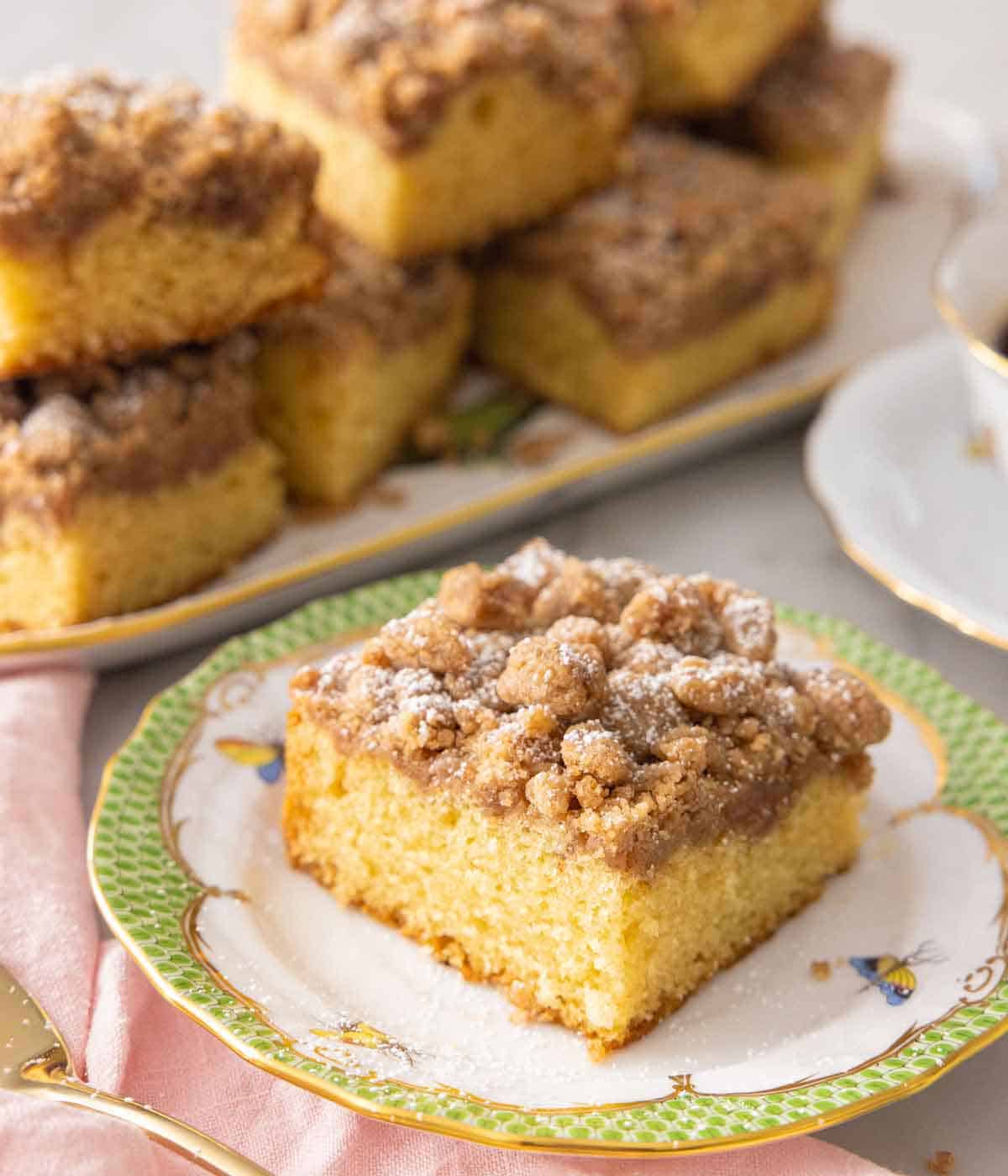 A slice of crumb cake on a plate in from of a platter of cakes.