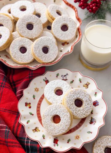 A plate with three linzer cookies by a mug of milk and a platter of cookies.