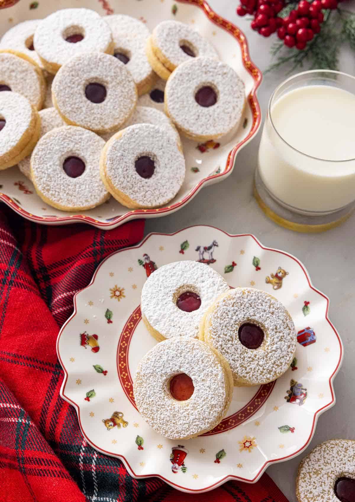 A plate with three linzer cookies by a mug of milk and a platter of cookies.