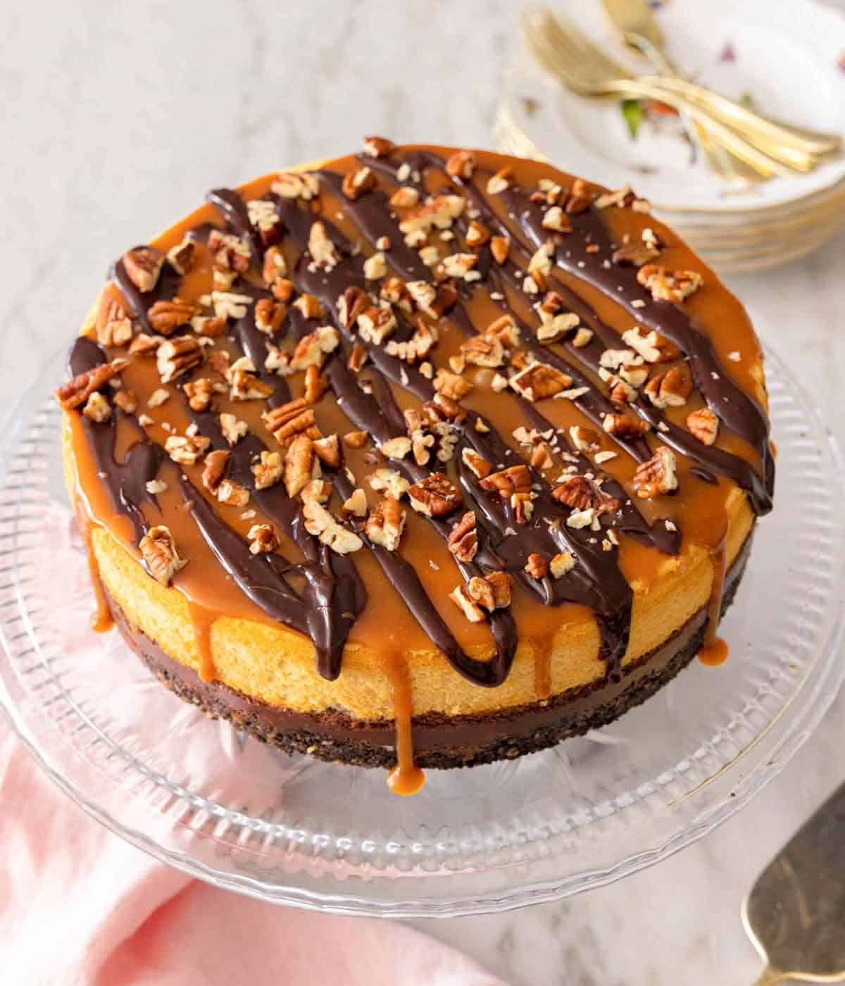 A turtle cheesecake on a cake pan with pecan, caramel, and chocolate on top.