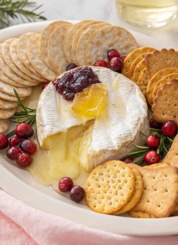 A platter with baked brie cut open with crackers around it and garnishes on top.