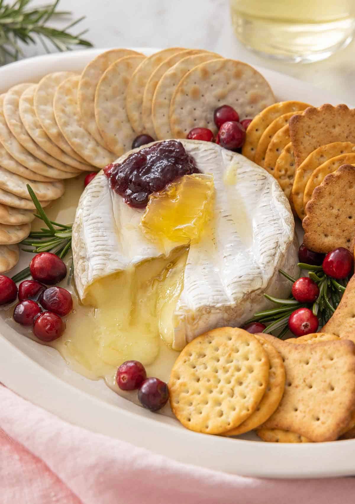 A platter with baked brie cut open with crackers around it and garnishes on top.
