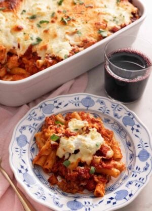 A plate with a serving of baked ziti by a glass of wine and baking dish.
