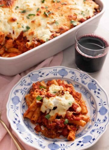 A plate with a serving of baked ziti by a glass of wine and baking dish.