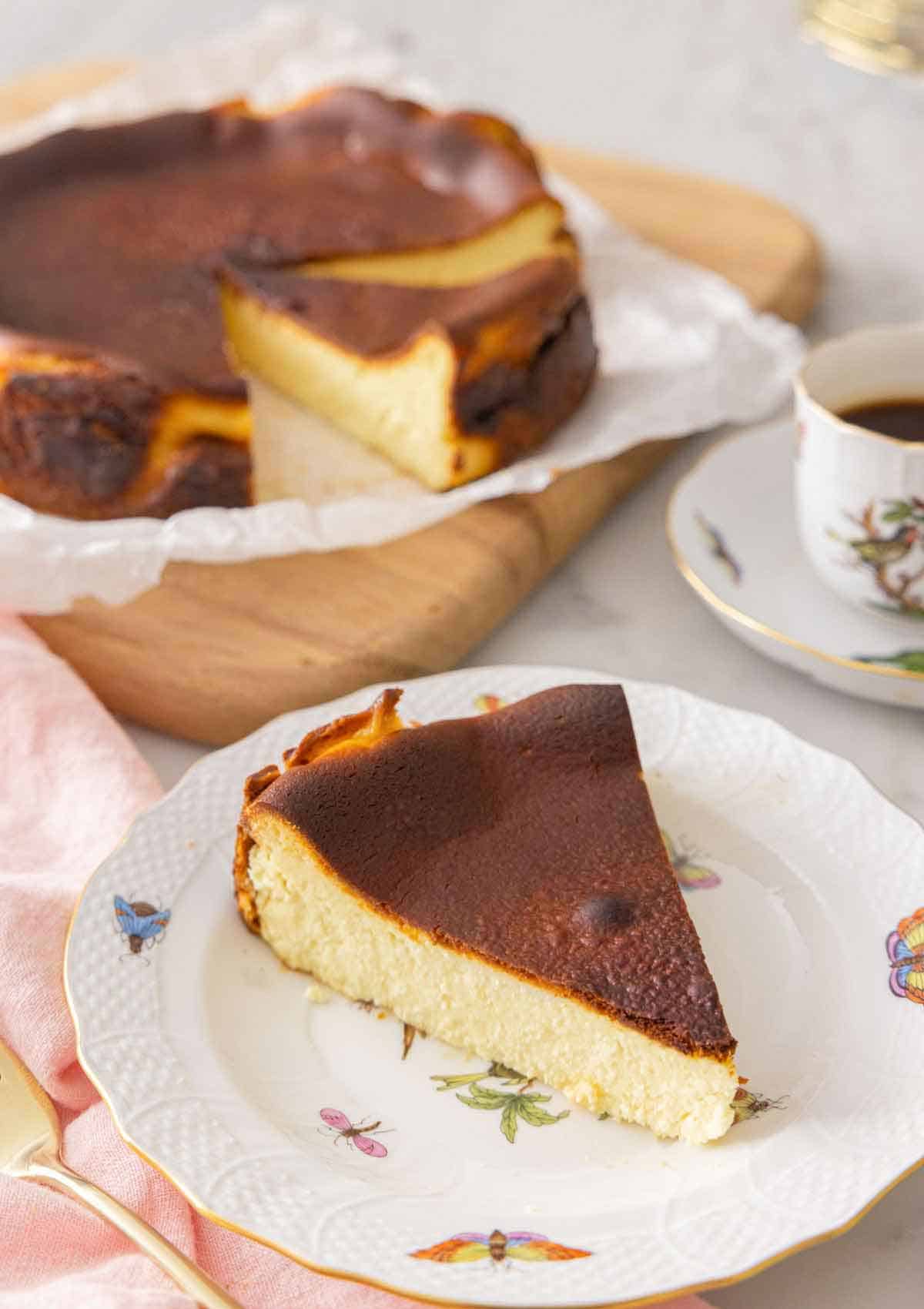 A slice of basque cheesecake on a plate in front the rest of the cake.