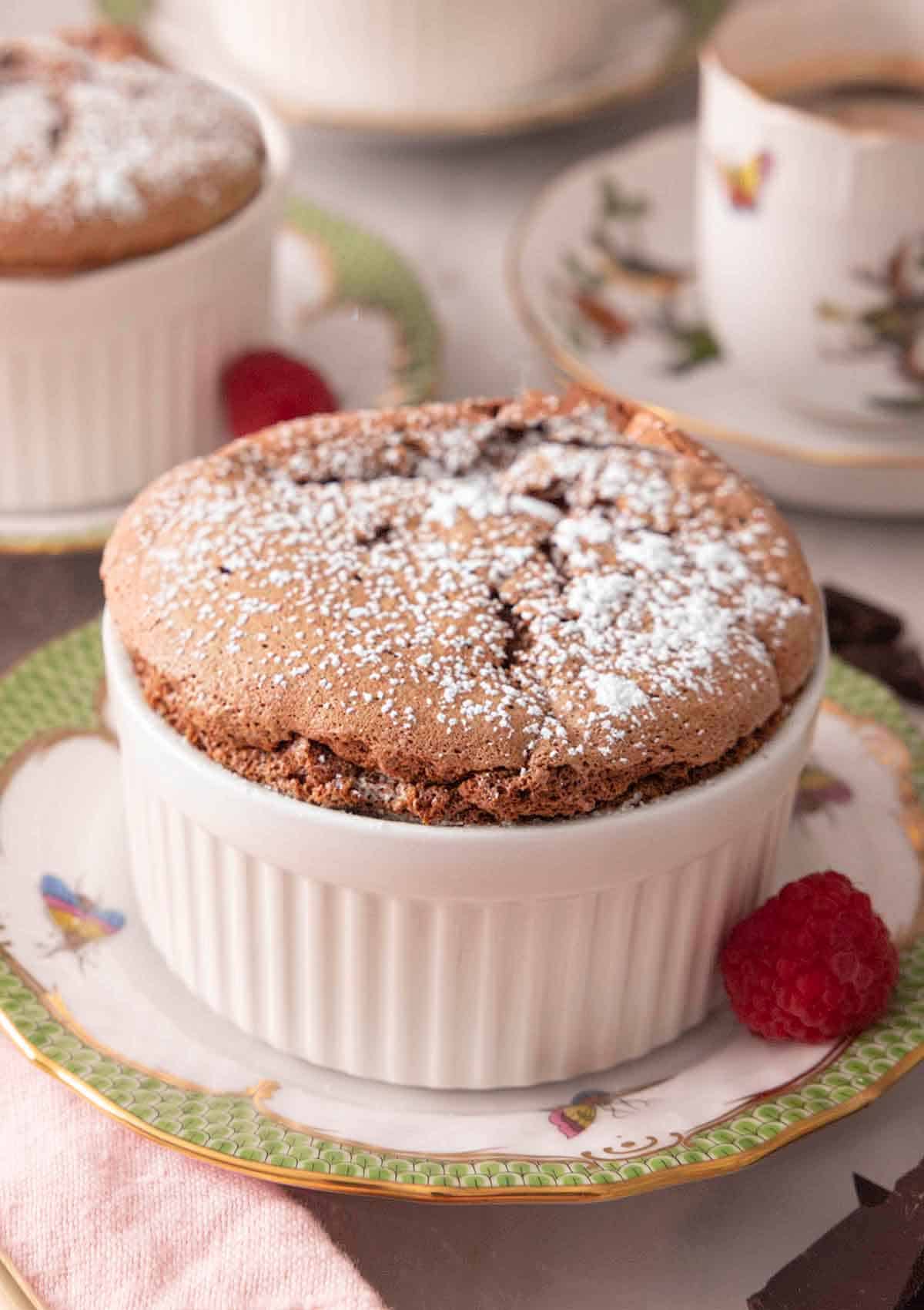 A ramekin of chocolate souffle with powdered sugar on top with a raspberry beside it.