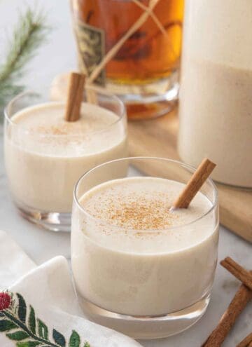 Two glasses of coquito with cinnamon sprinkles on top and a cinnamon stick garnish.