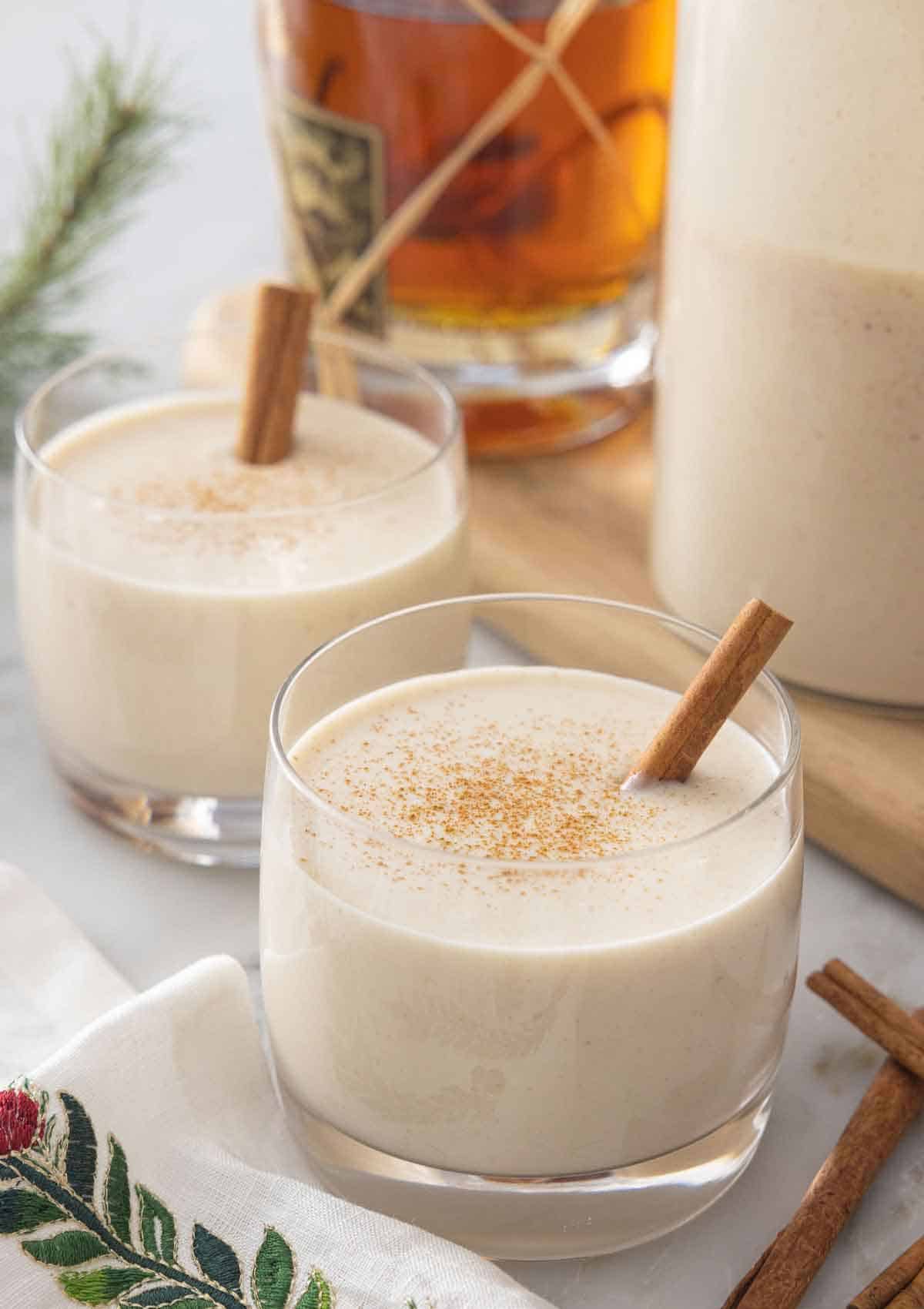Two glasses of coquito with cinnamon sprinkles on top and a cinnamon stick garnish.