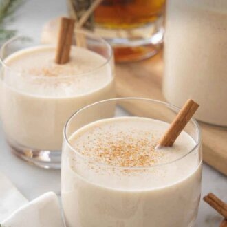 Pinterest graphic of two glasses of coquito with a bottle of rum in the back.