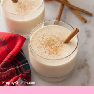 Pinterest graphic of two glasses of coquito with cinnamon sticks inside and on the counter.