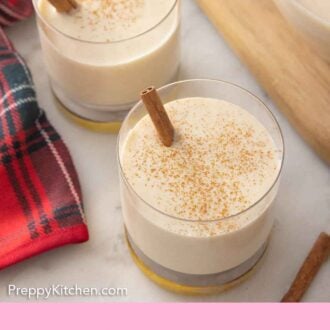 Pinterest graphic of an overhead view of two glasses of coquito with cinnamon powder and sticks.