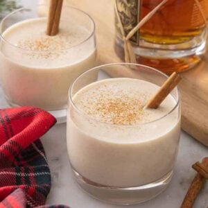 Two glasses of coquito with a bottle of rum in the background with a red linen in front.