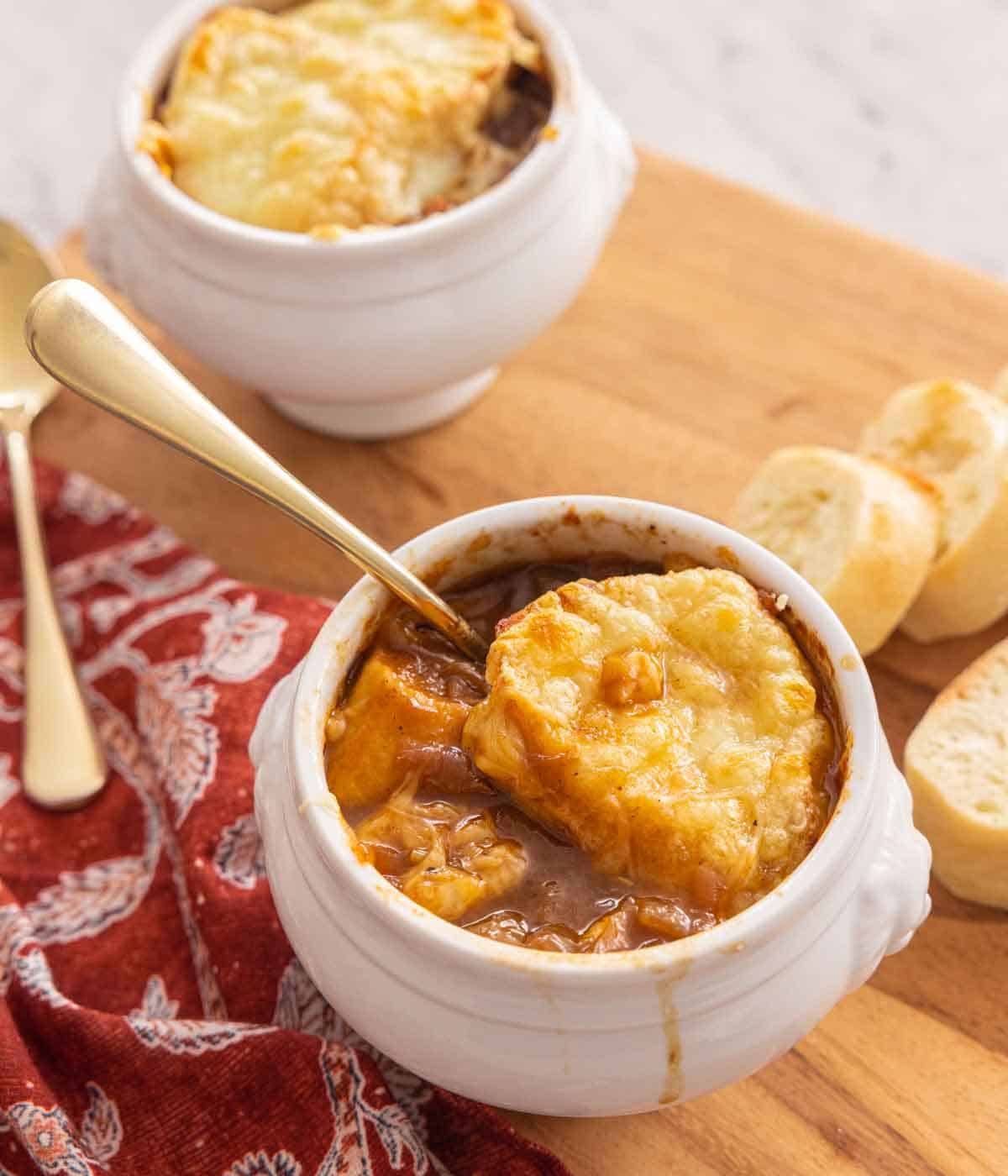 A bowl of French onion soup with a spoon inserted and sliced baguettes in the background.